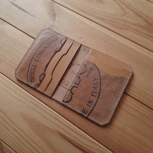 May include: Open tan leather wallet with multiple card slots. The wallet features black text that reads "SHELL CORDOVAN" and "CADO 1982 MADE IN ITALY". The wallet is open, revealing the interior card slots and a lighter tan leather lining.