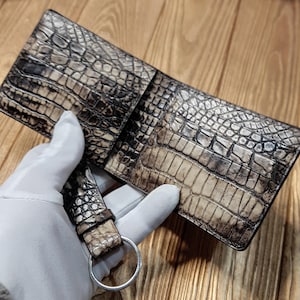 May include: A bi-fold wallet crafted from textured, light brown and dark grey crocodile leather. The wallet is open, showing the inside. A matching leather strap with a silver ring is attached.