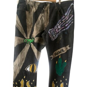 May include: Black denim jeans with hand-painted designs. The designs include a green lizard, a yellow eye, flames, a green dinosaur paw print, and a purple comet with the word "RAWR" on a banner.