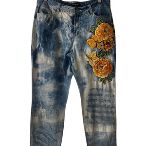 Peut inclure: Jean en denim bleu clair avec un effet tie-dye et une broderie florale jaune sur la jambe droite.