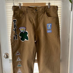 May include: Brown work pants with patches, including a green clover with the text "TRY IT" and a blue patch with the text "De-Bafflart 100+".