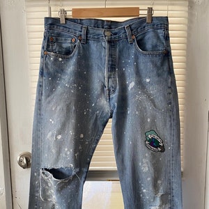 Puede incluir: Vaqueros de mezclilla azul desgastados con salpicaduras de pintura blanca y un parche en la pierna derecha que dice "Estuve perdido una vez, pero ahora me encontré".