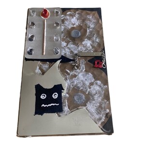 May include: A collage of mixed media art on a brown paper background. The collage includes a black fabric with a white cartoon face, a silver chain with a silver tag that says "CHLOE IS BACK", a red and white match, and a blister pack of pills.