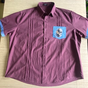 May include: A maroon short-sleeved button-down shirt with a blue pocket featuring a floral skull design. The shirt has decorative stitching on the front.