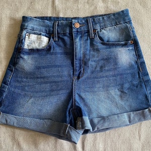 May include: A pair of blue denim shorts with a cuffed hem. The shorts have a small white pocket on the front left side.