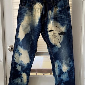 Peut inclure: Un jean en denim bleu délavé avec des éclaboussures de javel blanches et un texte noir qui dit "You see a wealthy you always were".