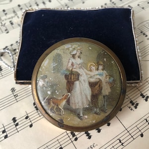 Op de afbeelding: Een vintage messing compact spiegel met een geschilderd tafereel van een vrouw en twee kinderen in een tuin. De spiegel is omgeven door een decoratief metalen frame en heeft een blauwe fluwelen koffer.