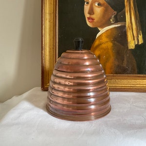 Könnte beinhalten: Ein kupferner Bienenstockbehälter mit einem schwarzen Knopf auf dem Deckel. Der Behälter hat eine glänzende, metallische Oberfläche und besteht aus mehreren Schichten.