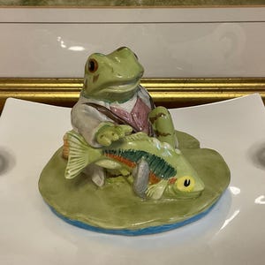 Op de afbeelding: Een keramieken beeldje van een kikker in een jas en vest, met een vis. De kikker is groen met bruine ogen en de vis heeft kleurrijke markeringen. Het beeldje staat op een groene waterlelie met een blauwe basis.