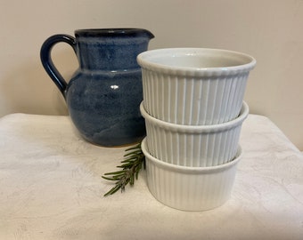 May include: A blue ceramic pitcher with a handle and a stack of three white, fluted ramekins. A sprig of rosemary is placed in front of the ramekins. The pitcher and ramekins are on a white tablecloth.
