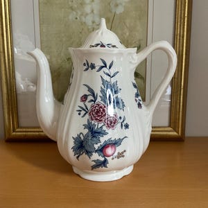 Könnte beinhalten: Weiße Keramik-Teekanne mit Deckel, verziert mit einem floralen Muster in Blau, Rosa und Grün. Die Teekanne hat einen gebogenen Ausguss und einen großen Griff. Im Hintergrund befindet sich ein gerahmtes Bild.