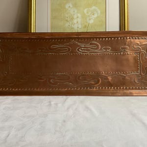 May include: A rectangular copper tray with decorative embossed scrollwork and dotted borders. The tray has a polished finish and a central recessed area. The overall color is a warm, reddish-brown. The tray is likely used for serving or display.