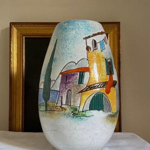 May include: A white ceramic vase with a hand-painted design of a small town with colorful buildings and a tree. The vase has a speckled glaze and a rounded shape.