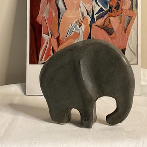 May include: A dark grey ceramic elephant figurine with a smooth, rounded surface. The elephant is standing on a white surface.