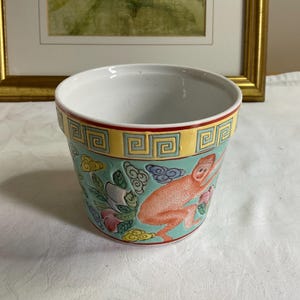 May include: A ceramic planter with a white interior and a colourful exterior. The exterior features a light teal background with a design of a monkey, flowers, and clouds. The rim has a yellow and red border with a geometric pattern.