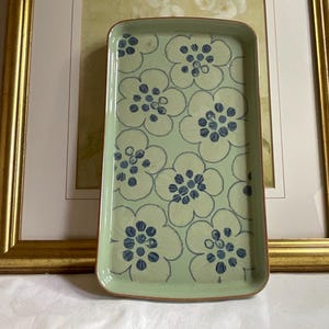 May include: Rectangular ceramic serving tray with a light green background and a repeating floral pattern in blue. The tray has rounded corners and a brown rim. The tray is displayed in front of a framed picture.