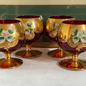 Può includere: Set di quattro calici in vetro rosso e oro con disegni floreali dipinti. I calici hanno una coppa larga e un gambo corto.