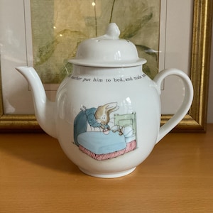 Può includere: Una teiera in ceramica bianca con coperchio, beccuccio e manico. La teiera presenta un'illustrazione colorata di un coniglio che mette a letto un altro coniglio, con testo sopra l'illustrazione. La teiera è appoggiata su una superficie di legno.