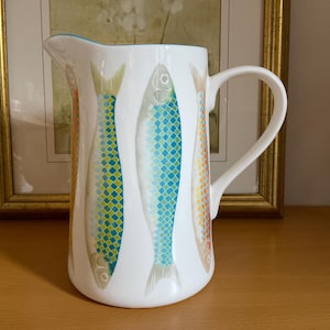 Op de afbeelding: Witte keramische kan met een handvat en een tuit, versierd met kleurrijke visillustraties. De vissen hebben blauwe, gele en oranje schubben. De kan heeft een lichtblauwe rand en staat op een houten oppervlak.