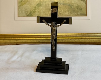 May include: A black wooden crucifix with a bronze-colored Jesus figure. The cross has brass-colored accents and sits on a tiered base. The background includes a framed painting.