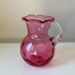 Könnte beinhalten: Eine rosa Glaskanne mit einem gewellten Rand und einem klaren Griff. Die Kanne hat eine strukturierte, blasenförmige Oberfläche.