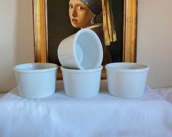 May include: Four white ceramic ramekins, perfect for baking individual desserts. The ramekins are cylindrical with a slightly flared rim. They are arranged on a white surface, with a painting in the background.