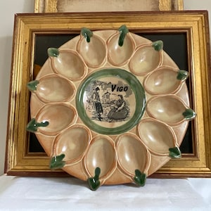 May include: A ceramic egg plate with 12 individual compartments, each with a green rim. The plate features a central design of two figures in traditional clothing, a building, and the word "Vigo".