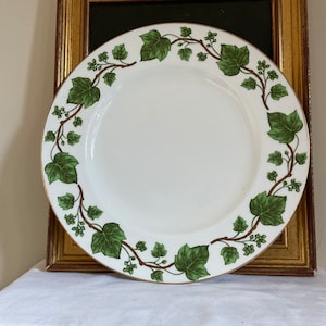 Może przedstawiać: Biały ceramiczny talerz z zielonym wzorem liści i jagód bluszczu na obrzeżu. Talerz ma złoty brzeg i jest ustawiony na tle w złotej ramie. Klasyczny element do serwowania.