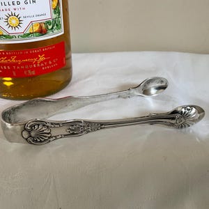 Peut inclure: Pinces à sucre en argent avec un design orné. Les pinces ont une extrémité arrondie en forme de cuillère.