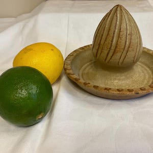 May include: A ceramic citrus juicer with a brown and tan glaze. The juicer has a ribbed top and a base with a scalloped edge. Two lemons and a lime are in the background.