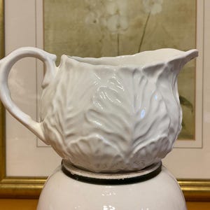 May include: White ceramic pitcher with a leaf-like textured design. The pitcher has a curved handle and a spout for pouring. It is sitting on top of a white ceramic base with a black band. The background includes a framed painting.