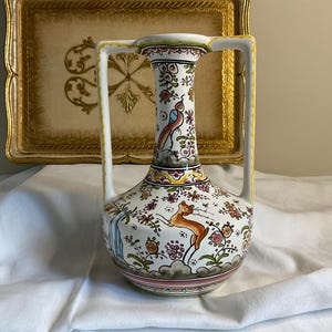 May include: A white ceramic vase with two handles, decorated with colourful floral and animal motifs. The vase features a bird, a deer, and a variety of flowers in shades of pink, blue, green, and yellow.