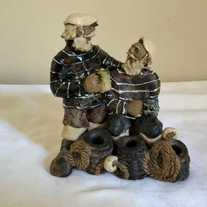 May include: A ceramic figurine depicting two bearded figures wearing cream-colored caps and dark brown striped sweaters. The figures are positioned behind three dark brown, textured cylindrical objects wrapped in rope, with one figure holding a small mug.