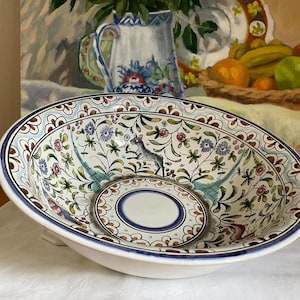 Könnte beinhalten: Eine runde Keramikschale mit handbemaltem Design, bestehend aus einem zentralen blauen Kreis, umgeben von floralen Mustern, grünem Blattwerk und einer kleinen grauen Hirschfigur. Der Rand ist mit einem braun-blauen Wellenmuster verziert.