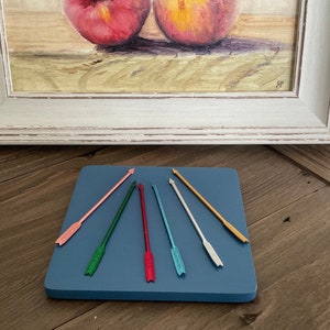 Op de afbeelding: Zeven kleurrijke pijlen met metallic afwerkingen, gerangschikt in een stervorm op een blauw vierkant oppervlak.
