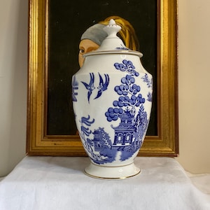 May include: A white porcelain vase with a blue and white willow pattern. The vase has a lid and is decorated with a pagoda, trees, and birds.