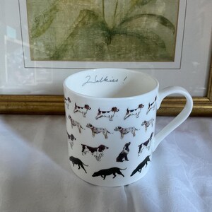 Könnte beinhalten: Weiße Keramiktasse mit Illustrationen verschiedener Hunde in Schwarz, Braun und Weiß. Die Tasse hat einen gebogenen Henkel und die Worte "2 Walkies!" sind auf dem Rand geschrieben. Die Tasse steht vor einem gerahmten Gemälde.