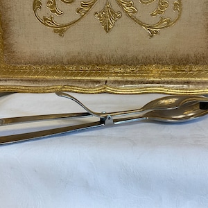 Könnte beinhalten: Silberfarbene Salat-Zange mit einem federbelasteten Griff und zwei löffelartigen Enden.