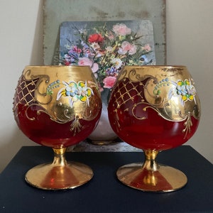 以下が含まれることがあります： 金色の縁取りと手描きの花柄が施された赤いガラス製のゴブレットが2つ。ゴブレットは、広口のボウルと短いステムを持っています。