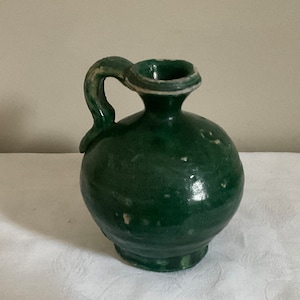 Könnte beinhalten: Ein kleiner, grüner Keramik Krug mit rundem Körper und einem gebogenen Henkel. Der Krug hat einen weißen Rand und eine leicht strukturierte Oberfläche.