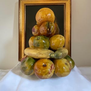 Può includere: Natura morta incorniciata con una piramide di frutta in ceramica. La composizione comprende arance, mele, pere e un grande melone, tutti con finiture lucide. La cornice è dorata e lo sfondo è scuro.