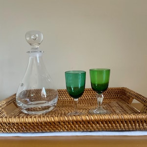 May include: A clear glass decanter with a spherical stopper and two small green glass goblets with clear stems, all arranged on a rectangular woven rattan tray with integrated handles and a light brown finish.