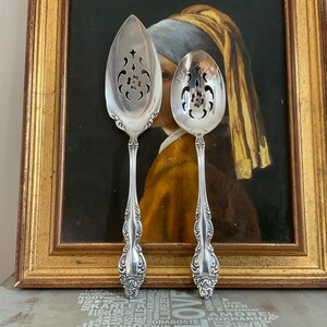 Puede incluir: Dos cucharas de servir plateadas con mangos ornamentados y cuencos calados. Las cucharas se exhiben frente a una pintura enmarcada con un marco dorado. Las cucharas tienen un diseño floral decorativo en los mangos.