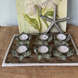 Może przedstawiać: Zestaw sześciu przezroczystych szklanych świeczników w kształcie gwiazdy z białymi świecami na drewnianej tacy z białą obwódką. Taca ma falisty wzór.