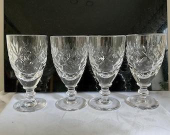 May include: Four clear crystal wine glasses with a diamond and leaf pattern. The glasses have a stemmed base and a rounded bowl. The glasses are arranged in a row on a white surface.