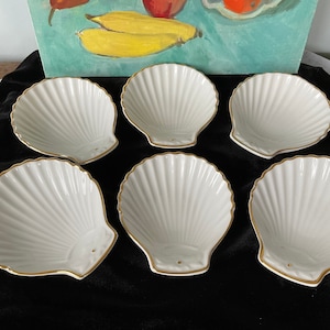May include: Six white porcelain seashell-shaped bowls with gold trim. The bowls are arranged in two rows of three on a black surface.