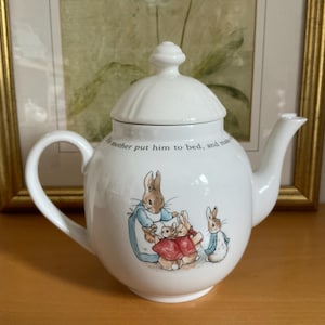 May include: White ceramic teapot with a lid, handle, and spout. The teapot features a colorful illustration of rabbits in clothing. Text on the teapot reads, "mother put him to bed, and made". The teapot is on a wooden surface.
