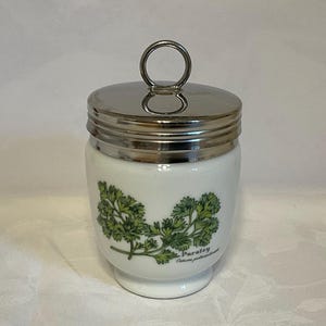 May include: A white ceramic jar with a shiny silver metal lid featuring a circular handle. The jar displays a green botanical illustration of parsley leaves and the text Parsley Carum petroselinum printed on the front surface of the container.