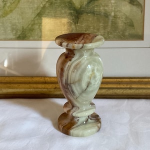 May include: A small, decorative vase made of onyx. The vase features a bulbous body with a flared top and base. The onyx has a swirling pattern of brown, tan, and cream colors. The vase is approximately 5 inches tall.