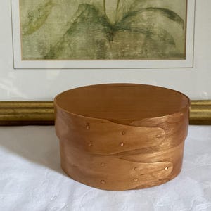 Könnte beinhalten: Eine runde Holzkiste im Shaker-Stil mit Deckel. Die Kiste ist hellbraun mit natürlicher Holzmaserung. Die Kiste hat einen Durchmesser von ca. 13 cm und ist 8 cm hoch. Die Kiste steht auf einer weißen Oberfläche.